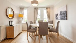 A dining area with a round table and six chairs, a wooden cabinet, a mirror, and a lamp. - Abbey Villa (No 5) (St Andrews)