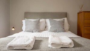 En pænt redt seng med hvide sengetøj og to puder med mønster, en sengebordlampe og et træskab.