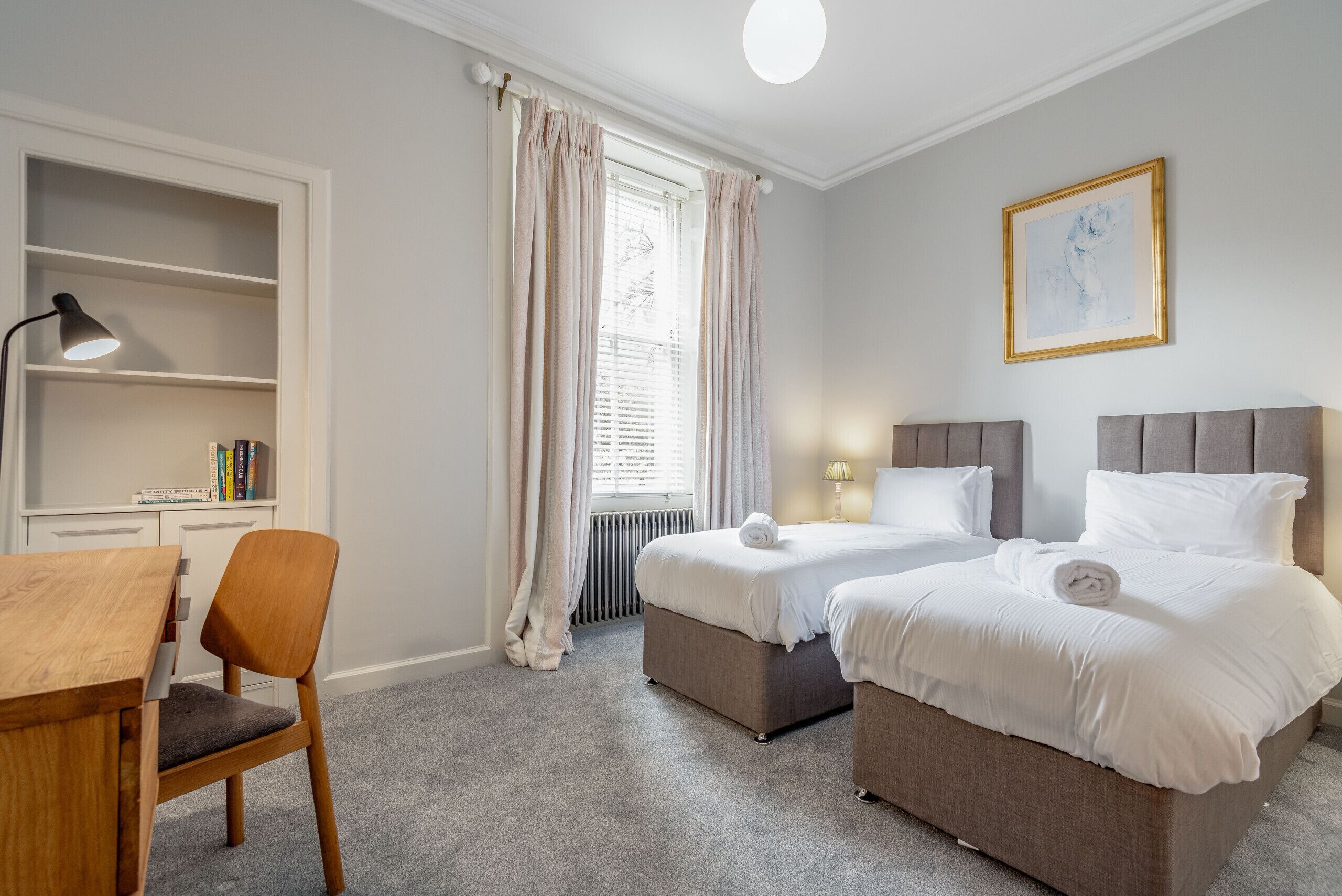 Una habitació d'hotel amb dos llits, un escriptori, una cadira, una estanteria i una finestra amb cortines.