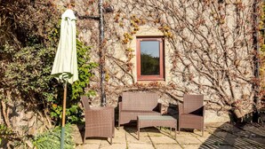A patio with wicker furniture and a table, a covered umbrella, and a window with a view of trees. - Old Mill Cottage (Dunino)