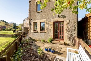 A stone building with a wooden deck, a red door, and a garden with potted plants. - Old Mill Cottage (Dunino)