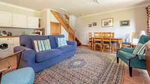 A living room with a sofa, chairs, a dining table, a staircase, and a kitchen area. - Old Mill Cottage (Dunino)