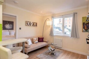 Un salon moderne avec un canapé, une table basse et un lampadaire. Une fenêtre offre une vue sur les bâtiments environnants.