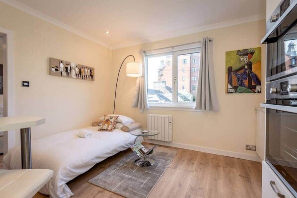 Une chambre à coucher avec un lit, une petite table, un radiateur, une fenêtre avec des rideaux et un tableau accroché au mur.
