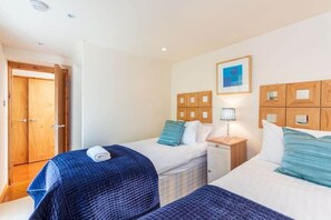 A bedroom with two beds, wooden headboards, a small bedside table with a lamp, and a door to the left.
