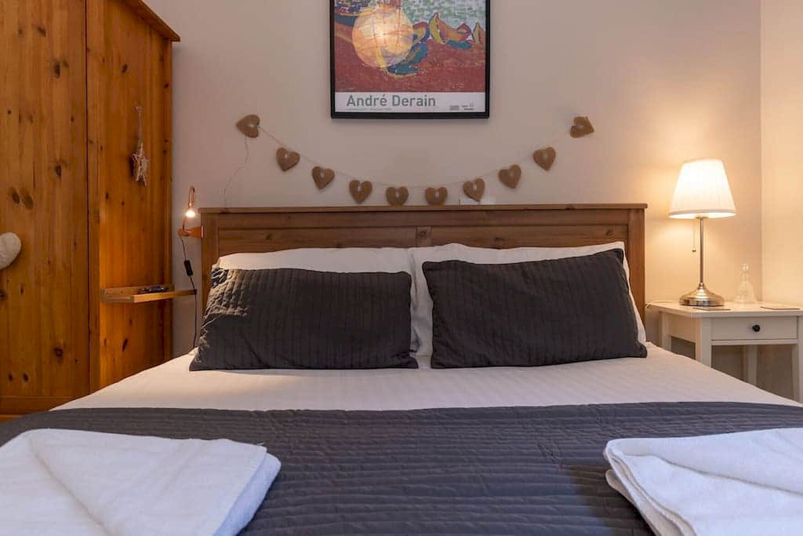 A neatly made bed with two pillows, a wooden headboard, a wall-mounted picture, a heart-shaped decoration, and a bedside lamp.