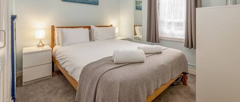 A neatly arranged bedroom with a bed, bedside tables, a nightstand, a mirror, and a window with curtains.