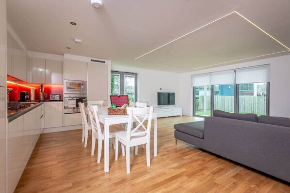 Una cocina moderna con armarios blancos, una mesa de comedor con sillas, un sofá gris y un televisor.