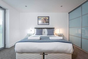 A modern bedroom with a large bed, two bedside tables with lamps, a framed picture on the wall, and a sliding glass door to the right. - Square Green | Garden Apartment With Putting Gree (St Andrews)