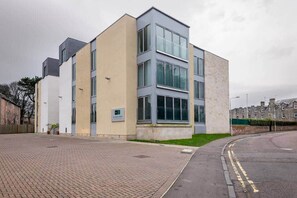 A modern building with a flat roof, large windows, and a brick-paved entrance. - Square Green | Garden Apartment With Putting Gree (St Andrews)
