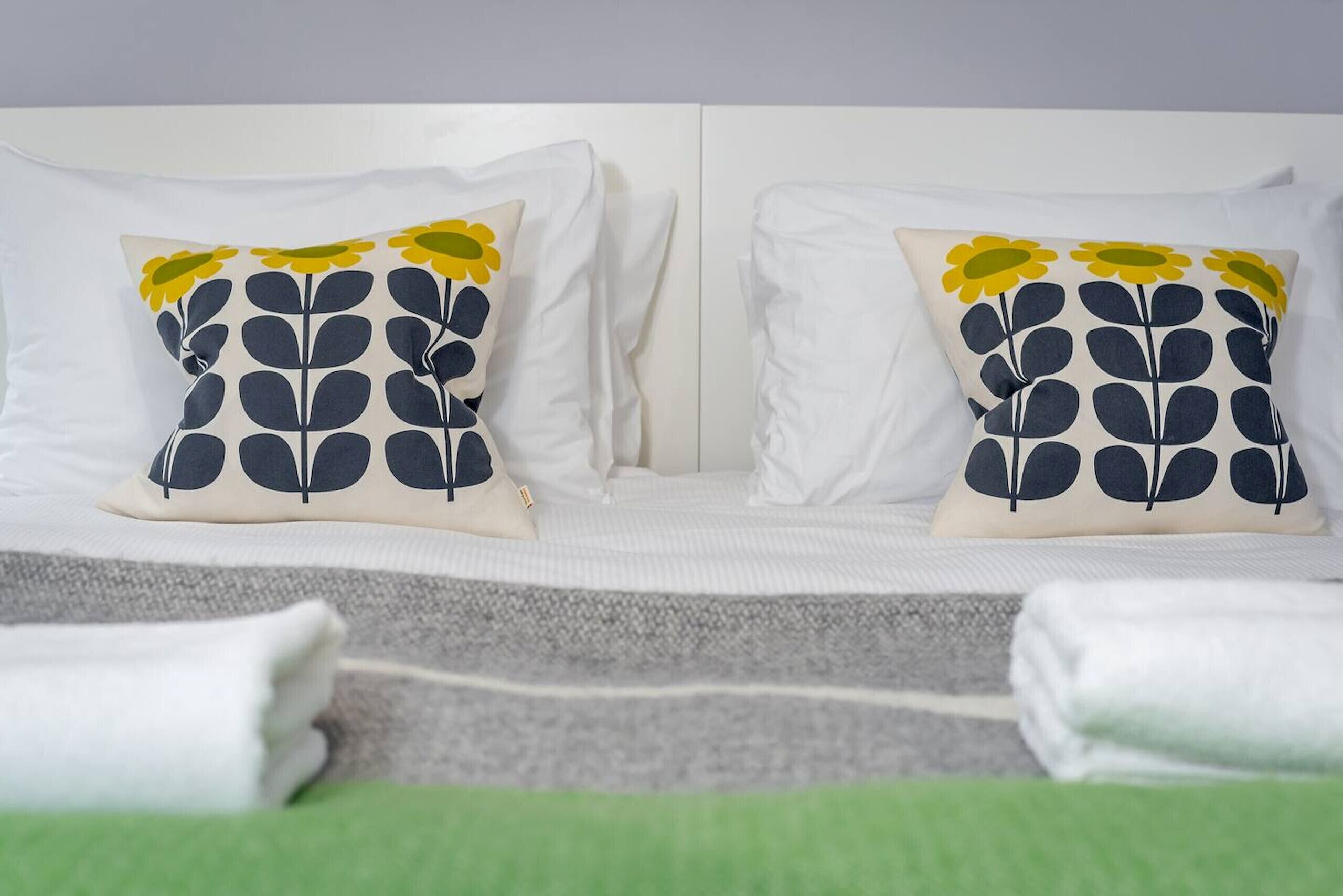 A bed with white bedding, two decorative pillows with a leaf pattern, and neatly folded white towels.