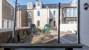Une cour avec un banc, deux chaises et une petite table. On y voit des bâtiments avec des fenêtres et un escalier menant à un étage supérieur.