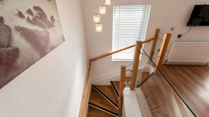Un escalier moderne avec des marches en bois et une rampe en verre, un téléviseur fixé au mur et une fenêtre avec des stores.