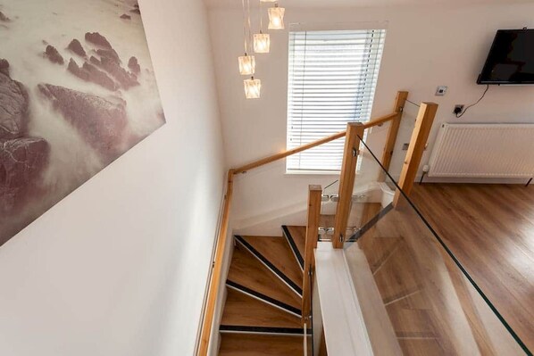 A modern staircase with wooden steps and glass railings, a wall-mounted TV, and a window with blinds. - Old Net Store | Anstruther (Anstruther)