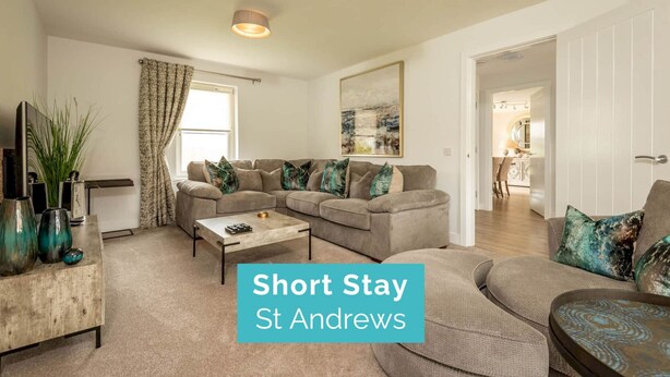 A modern living room with a sectional sofa, a coffee table, and a TV. There is a painting on the wall and a plant on the left. The text "Short Stay St Andrews" is visible. - Village Green | Kingsbarns (Kingsbarns)