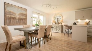 A modern kitchen with a dining area, featuring a large island, pendant lighting, and a view of the living area. - Village Green | Kingsbarns (Kingsbarns)