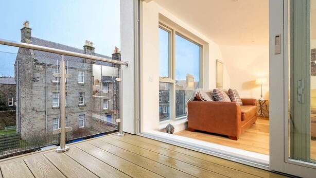 A balcony with a view of a stone building, a wooden floor, and a brown sofa with cushions.