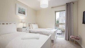 A hotel room with two beds, a window with curtains, a small stool, and a television on the wall.