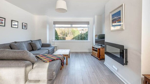 Una sala de estar moderna con un sofá gris, una mesa de centro de madera, un televisor de pantalla plana y un amplio ventanal con vistas a la vegetación.