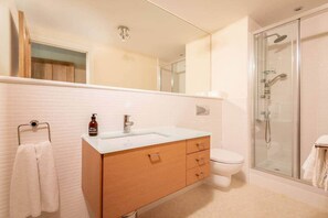 Ein Badezimmer mit weiß gefliesten Wänden, einer Glasdusche, einem Holzkosmetikschrank mit Waschbecken und einer Toilette.
