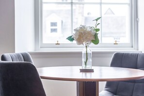 A round dining table with a vase of flowers and a book, set against a window with a view of buildings.