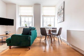 A modern living room with a sofa, a dining table with chairs, a flat-screen TV, and large windows with a view.