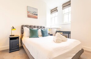 A neatly arranged bedroom with a bed, two bedside tables, lamps, a painting, and a window with curtains.