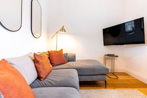 A modern living room with a grey sofa set, a floor lamp, a flat-screen TV mounted on the wall, and a small side table.