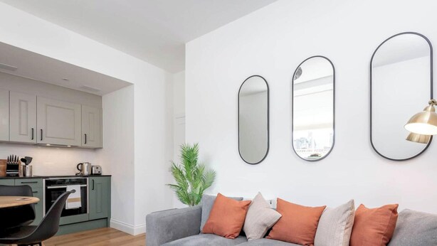 Una sala moderna con un sofá gris, cojines naranjas y una lámpara de pie. Se ve en el fondo una cocina con armarios blancos y un comedor con mesa y sillas.