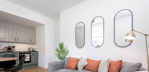 Uma sala de estar moderna com um sofá cinzento, almofadas laranja e um abajur. Uma cozinha com armários brancos e uma área de refeições com mesa e cadeiras são visíveis ao fundo.