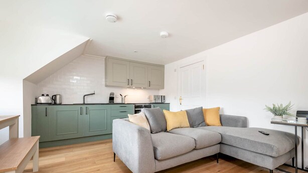 A modern kitchen with light green cabinets, a grey sofa with cushions, and a wooden dining table. - Skye Sands | Balgove Penthouse Residence (St Andrews)