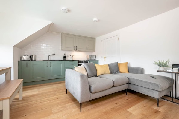 A modern kitchen with green cabinets, a grey sofa with cushions, and a wooden dining table.
