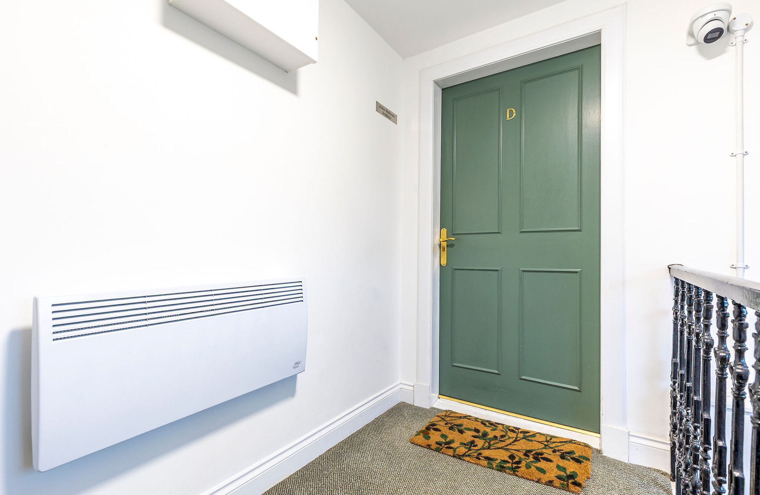 Una puerta verde con felpudo, un radiador blanco y una cámara de seguridad.