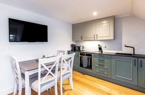 A modern kitchen with grey cabinets, a flat-screen TV mounted on the wall, and a dining area with a wooden table and white chairs.
