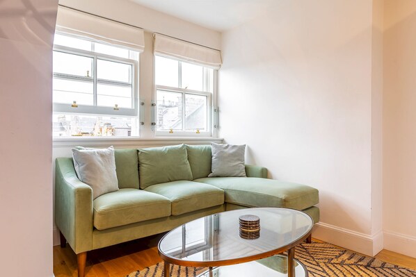 A room with a green sofa, a glass coffee table, and a window with white blinds.
