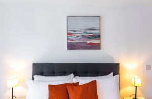 Une chambre à coucher comprenant un lit, des oreillers, une tête de lit, un tableau accroché au mur et deux lampes de chevet.