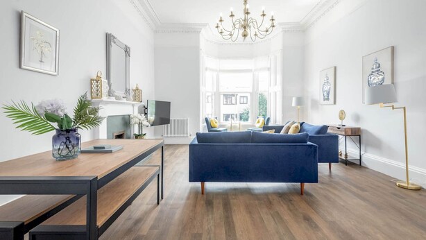 A modern living room with a blue sofa, a wooden dining table, and a chandelier. - Skye Sands | Alexandra Penthouse (No 8) (St Andrews)