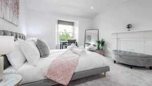 A modern bedroom with a large bed, a grey upholstered bench, a small table, and a potted plant. - Skye Sands | Alexandra Penthouse (No 8) (St Andrews)