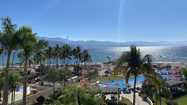 A resort with a pool area, palm trees, and a view of the ocean and mountains.