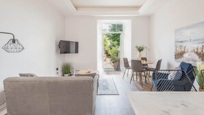 Ein modernes Wohnzimmer mit einer Couch, einem Essbereich mit Stühlen und einem Tisch, einem an der Wand befestigten Fernseher und einem großen Fenster mit Blick ins Freie.