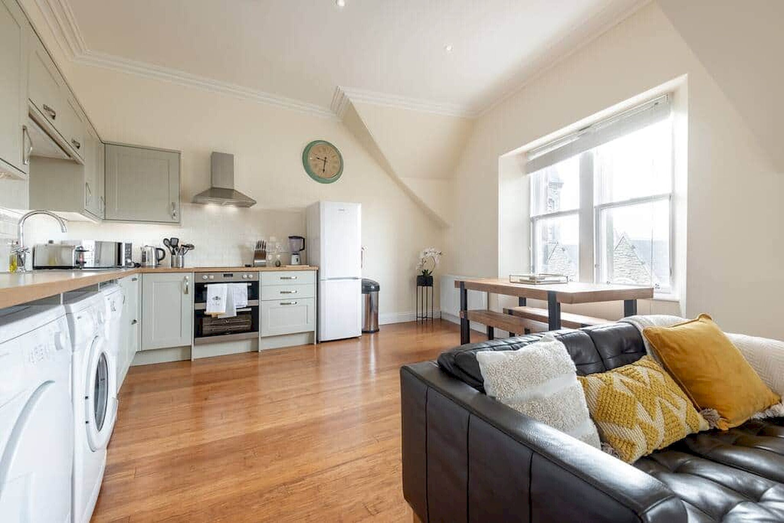 A modern kitchen with white cabinets, a built-in oven, and a coffee machine. There is a dining table with chairs and a black leather sofa with cushions.