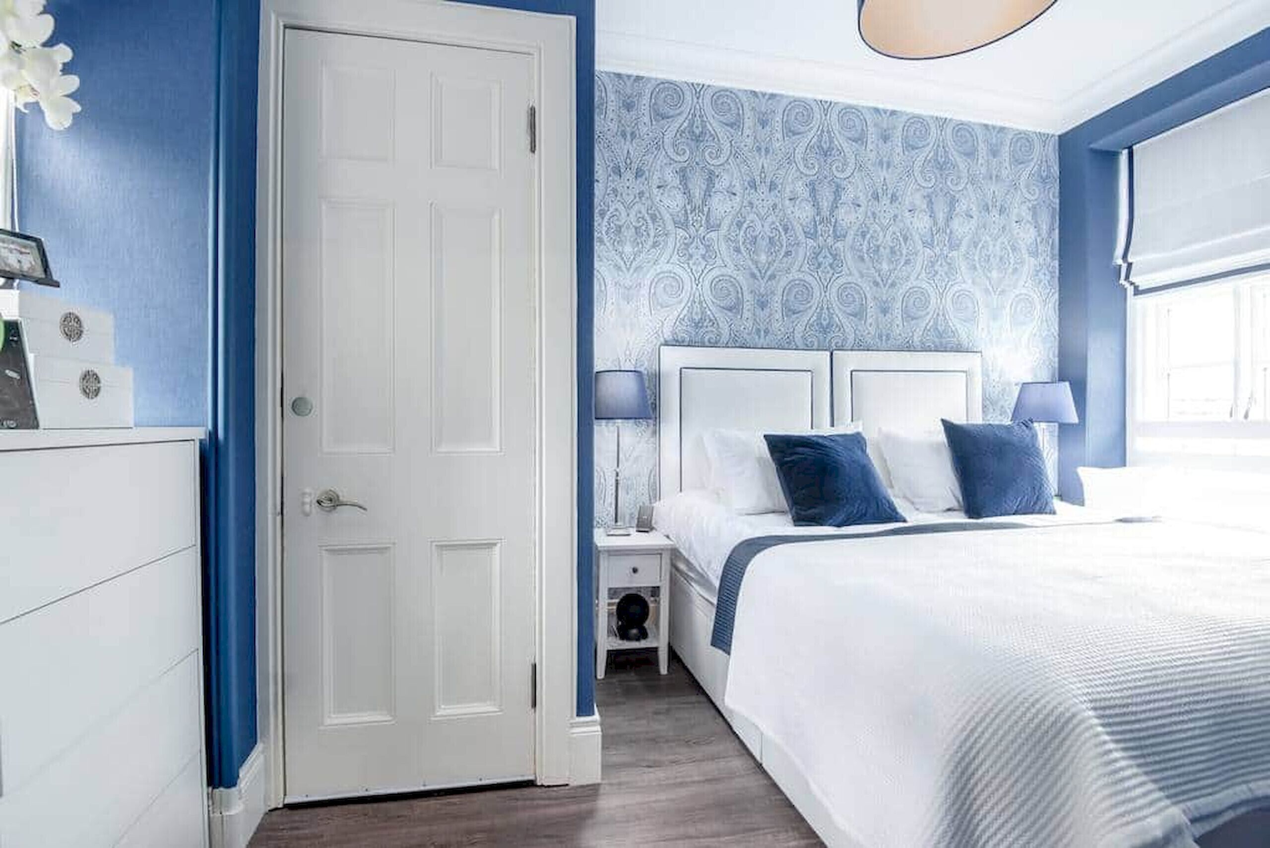 Un dormitorio con puerta blanca, cama con ropa de cama azul y blanca, mesita de noche con lámpara y pared con diseño.