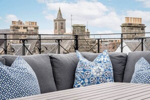Une terrasse sur le toit offrant une vue sur des bâtiments historiques, dont le clocher d’une église, ainsi que des sièges rembourrés de couleur grise agrémentés de coussins à motifs.