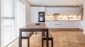 Une cuisine moderne avec des meubles blancs, une table en bois et une grande fenêtre équipée de stores.