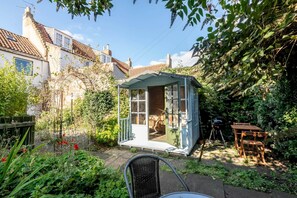 A small, enclosed garden area with a wooden table and chairs, surrounded by lush greenery and a house in the background.