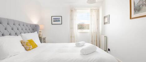 A neatly made bed with white linens and a single yellow pillow, a grey upholstered headboard, and a framed picture on the wall.