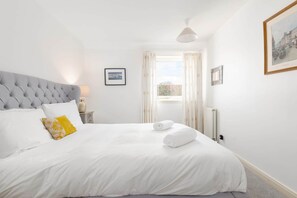 A neatly made bed with white linens and a single yellow pillow, a bedside table with a lamp, and a framed picture on the wall.
