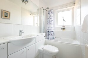 A modern bathroom with a white sink, toilet, and bathtub. There is a window with a curtain and a small bird decoration.
