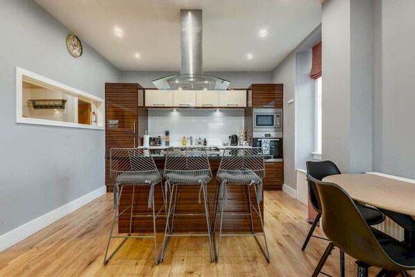 A modern kitchen with a central island, bar stools, a dining table, and a clock on the wall.
