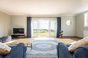 A living room with a television, seating, a large window with curtains, and a view of the outdoors. - Firthview | Crail (Anstruther)